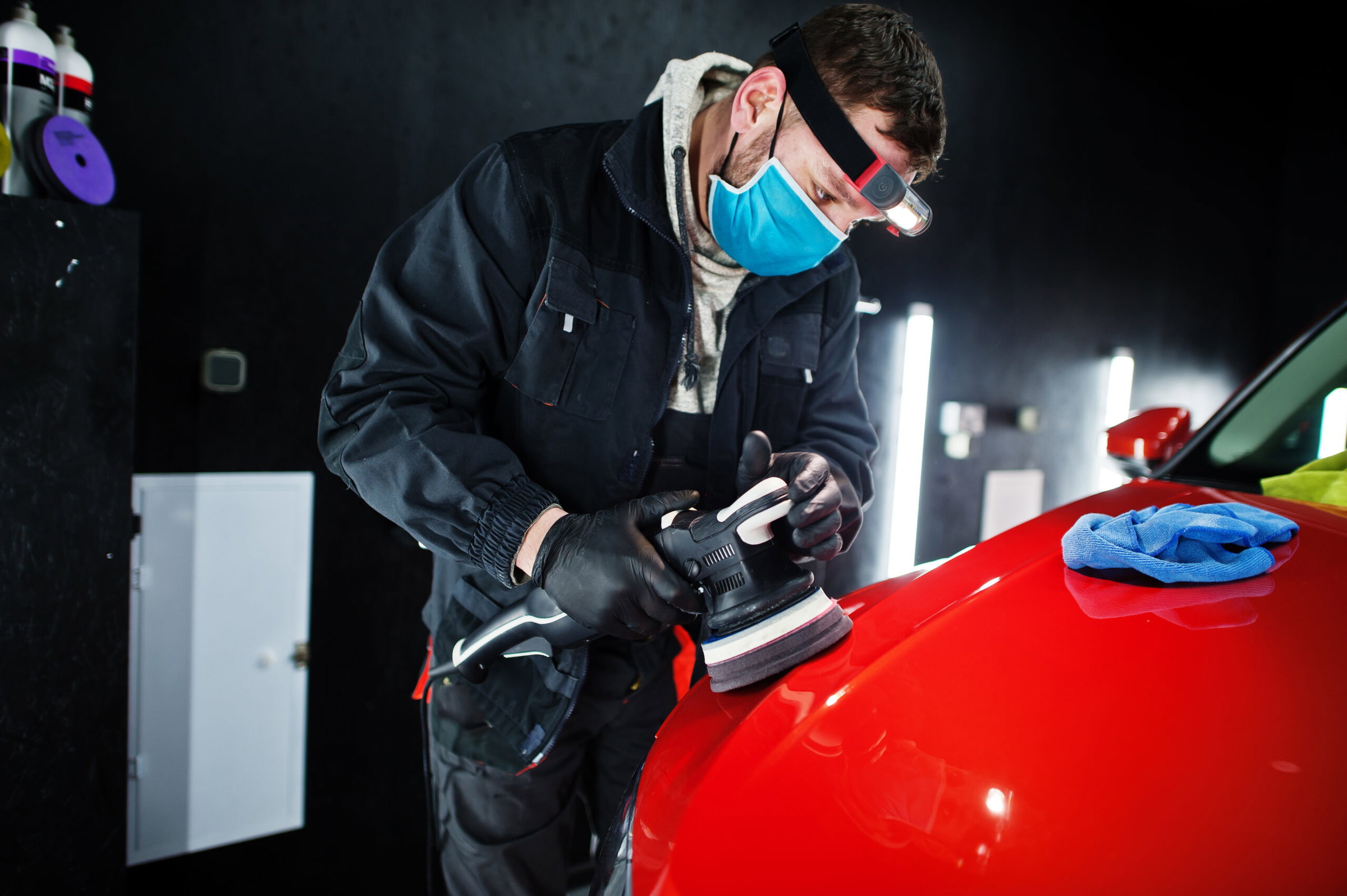 A man wearing a mask, gloves, and headlamp is polishing the hood of a red car with a power buffer in an auto body shop workshop.
