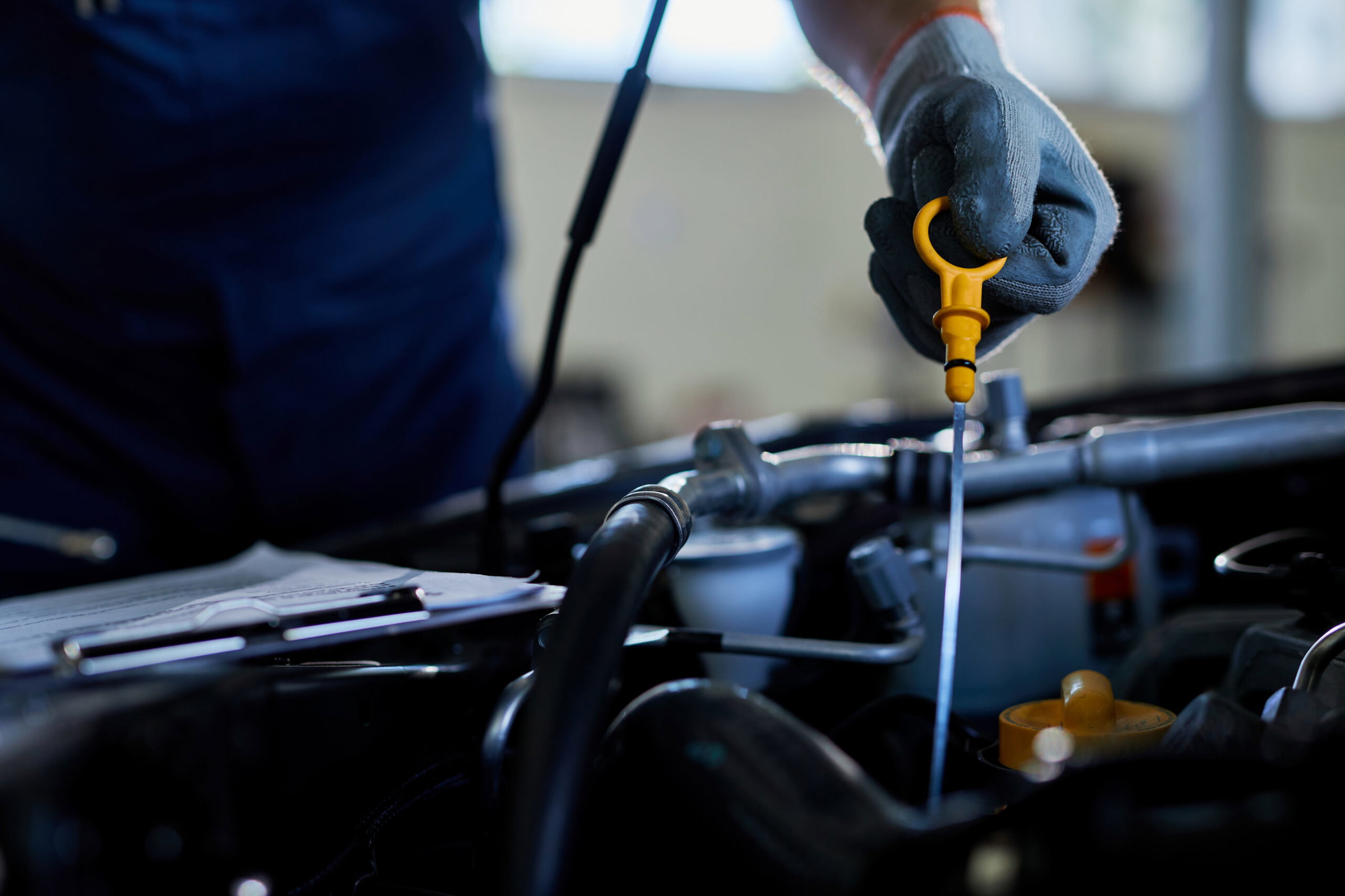 A person wearing gloves checks a car’s oil level by holding the dipstick over the engine bay at an auto repair shop. A clipboard is visible in the background. mechanical services