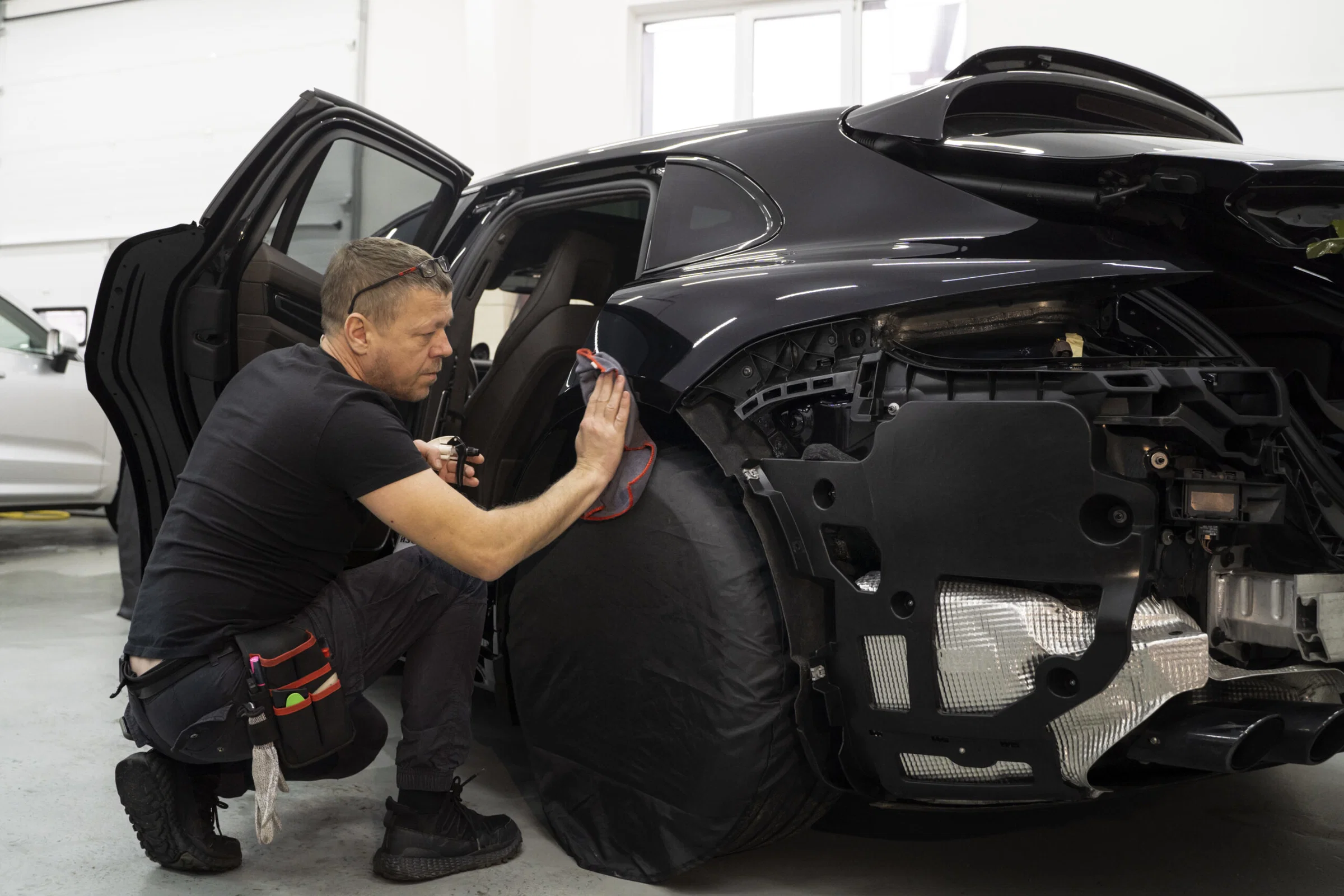A man kneels beside a black car with its front bumper removed, cleaning or inspecting the exposed wheel area in a Bay Area body shop. Collision repair